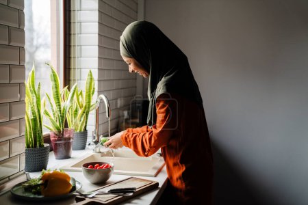 Foto de Vista lateral de la joven mujer tranquila en el hijab lavado de verduras en la acogedora cocina en casa - Imagen libre de derechos