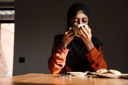 Téléchargez les photos : Jeune belle femme calme dans le hijab boire du thé avec des cookies - en image libre de droit