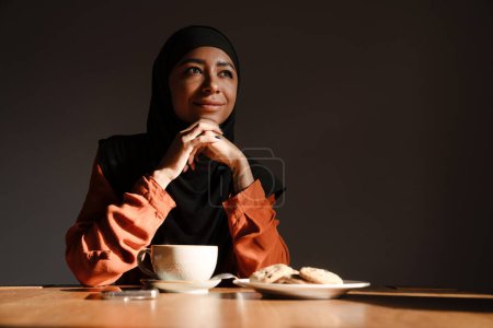 Téléchargez les photos : Jeune belle femme calme en hijab assise à la table avec une tasse de thé et des biscuits - en image libre de droit