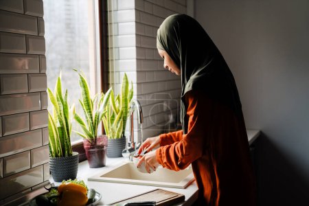Téléchargez les photos : Vue de côté de la jeune femme calme en hijab laver les légumes dans une cuisine confortable à la maison - en image libre de droit