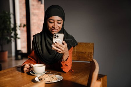 Foto de Joven hermosa mujer sonriente feliz en hijab con teléfono sentado en la mesa de la cocina con taza de té y galletas - Imagen libre de derechos