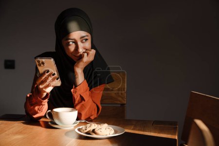 Téléchargez les photos : Jeune belle femme calme dans le hijab avec téléphone assis à la table de cuisine avec une tasse de thé et des biscuits soutenant sa tête et regardant vers la droite - en image libre de droit
