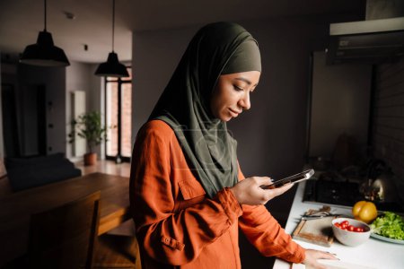 Foto de Joven mujer tranquila en hijab mirando su teléfono mientras está de pie junto a la propagación de verduras en la acogedora cocina en casa - Imagen libre de derechos