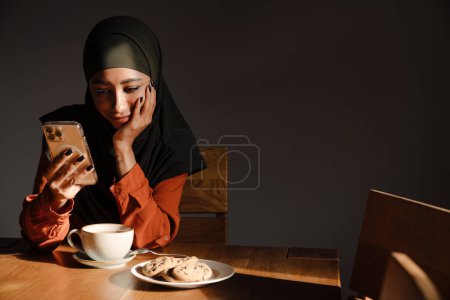 Téléchargez les photos : Jeune belle femme calme en hijab avec téléphone assis à la table de cuisine avec tasse de thé et biscuits - en image libre de droit