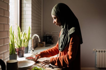 Photo for Sideview of young calm woman in hijab preparing vegetables in cozy kitchen at home - Royalty Free Image