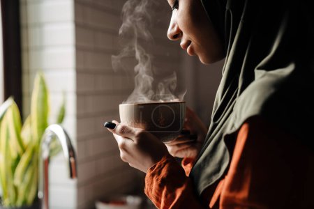 Téléchargez les photos : Jeune femme calme dans le hijab tenant une tasse de thé chaud devant la fenêtre dans une cuisine confortable à la maison - en image libre de droit