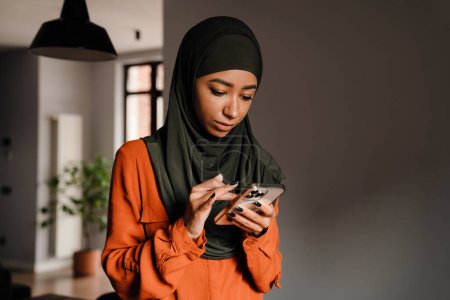 Foto de Joven mujer tranquila en hijab desplazando su teléfono en casa - Imagen libre de derechos