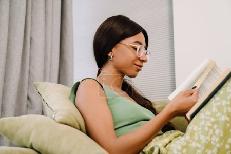 Foto de Joven mujer sonriente atractiva en gafas sentadas en el sofá y leyendo el libro en casa - Imagen libre de derechos