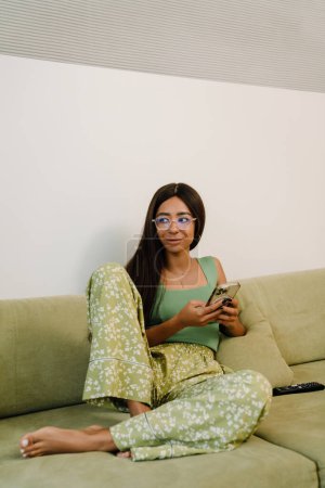Téléchargez les photos : Jeune belle femme souriante dans des lunettes assis sur le canapé avec téléphone regardant vers la gauche à la maison - en image libre de droit