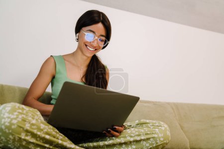 Téléchargez les photos : Jeune belle femme souriante dans des lunettes et des écouteurs travaillant avec un ordinateur portable tout en étant assis à la maison sur le canapé dans la pose de lotus - en image libre de droit