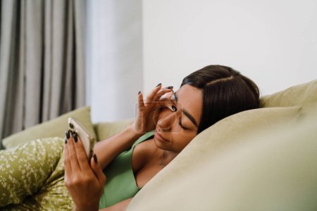 Téléchargez les photos : Jeune belle femme fatiguée avec téléphone frottant ses yeux couché sur le canapé à la maison - en image libre de droit