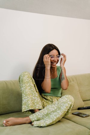 Foto de Joven atractiva feliz sonriente mujer ajustando gafas sentado en el sofá en casa - Imagen libre de derechos
