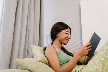 Téléchargez les photos : Jeune femme souriante attrayant dans des lunettes assis sur le canapé et la lecture du livre à la maison - en image libre de droit