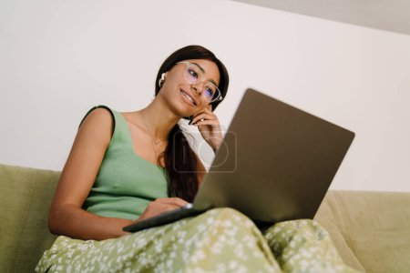 Téléchargez les photos : Jeune belle femme souriante dans des lunettes et écouteurs assis sur le canapé et travaillant avec un ordinateur portable à la maison - en image libre de droit