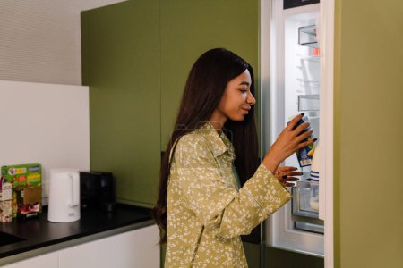 Téléchargez les photos : Jeune femme attrayante aux cheveux longs prenant un paquet de jus du réfrigérateur dans la cuisine à la maison - en image libre de droit