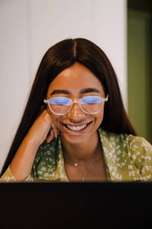 Foto de Retrato de joven hermosa mujer sonriente en gafas y blusa verde mirando en la pantalla del ordenador portátil y tocando su mejilla - Imagen libre de derechos