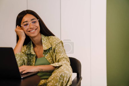 Foto de Joven hermosa mujer sonriente en gafas apoyando su cabeza y mirando a un lado sentado con el ordenador portátil en casa - Imagen libre de derechos