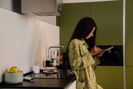 Téléchargez les photos : Jeune femme attrayante aux cheveux longs debout avec tablette dans la cuisine à la maison - en image libre de droit