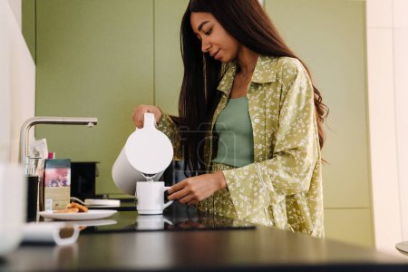 Téléchargez les photos : Jeune belle femme aux cheveux longs versant de l'eau chaude dans une tasse pour faire du thé / café dans une cuisine confortable à la maison - en image libre de droit