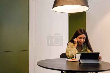 Téléchargez les photos : Jeune belle femme aux cheveux longs avec une tasse de café avec des cookies tablette défilante à la maison - en image libre de droit
