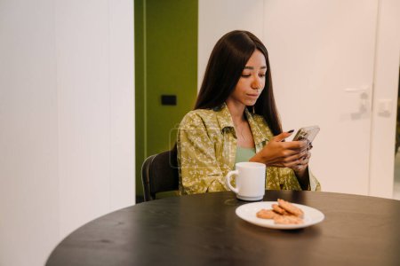 Photo for Young beautiful long-haired woman sitting at kitchen table with cup of coffee and cookies looking on her phone at home - Royalty Free Image