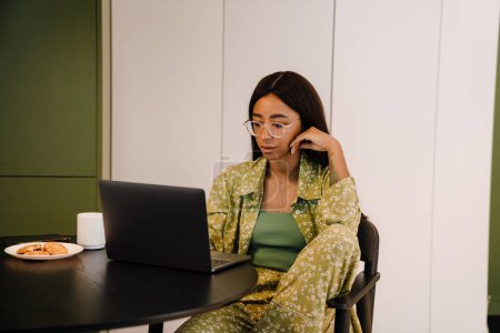 Foto de Joven hermosa mujer seria en gafas de trabajo en el ordenador portátil con taza de café y galletas en casa - Imagen libre de derechos