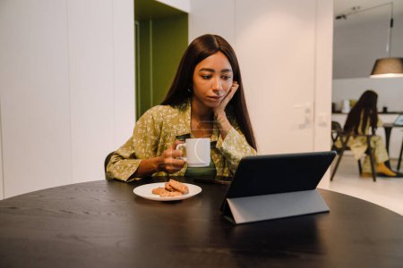 Foto de Joven hermosa mujer de pelo largo bebiendo café con galletas y viendo series en su tableta en casa - Imagen libre de derechos