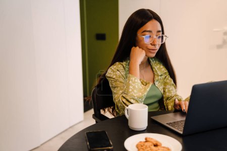 Téléchargez les photos : Jeune belle femme aux cheveux longs dans des lunettes de travail sur ordinateur portable avec tasse de café et biscuits à la maison - en image libre de droit