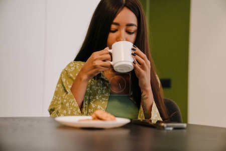 Foto de Joven hermosa mujer de pelo largo sentado en la mesa de la cocina beber café con galletas en casa - Imagen libre de derechos