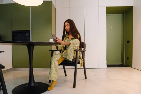 Téléchargez les photos : Jeune belle femme aux cheveux longs assise à la table de cuisine avec une tasse de café et regardant son téléphone à la maison - en image libre de droit