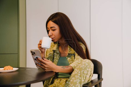Foto de Joven hermosa mujer de pelo largo sentada en la mesa de la cocina bebiendo café y mirando en su teléfono en casa - Imagen libre de derechos
