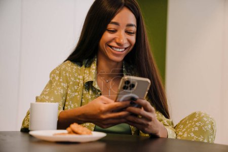 Téléchargez les photos : Jeune femme souriante aux cheveux longs assise avec une tasse de café et des biscuits regardant son téléphone à la maison - en image libre de droit