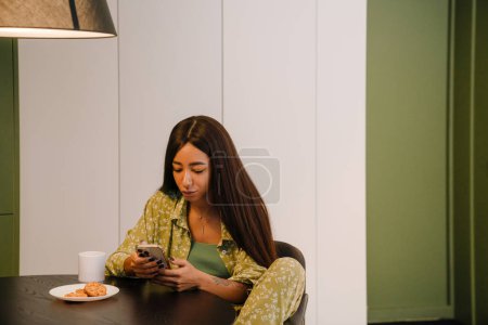 Téléchargez les photos : Jeune belle femme aux cheveux longs assise à la table de cuisine avec une tasse de café et des biscuits regardant son téléphone à la maison - en image libre de droit