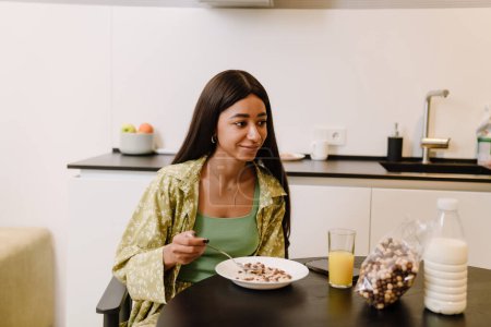 Foto de Joven hermosa calma mujer de pelo largo sentado a la mesa y comer hojuelas con leche en el desayuno en la acogedora cocina t casa - Imagen libre de derechos