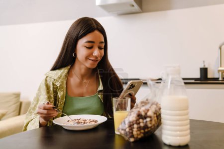 Téléchargez les photos : Jeune belle femme souriante aux cheveux longs regardant son téléphone tout en mangeant des flocons de lait au petit déjeuner dans une cuisine confortable à la maison - en image libre de droit