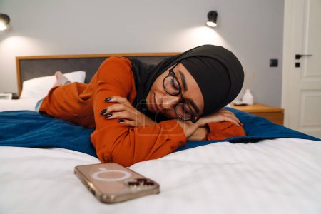 Téléchargez les photos : Jeune belle femme en hijab et lunettes dormant sur le lit avec téléphone - en image libre de droit