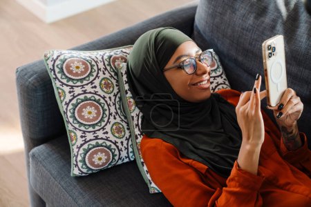 Foto de Joven hermosa mujer sonriente en hijab y gafas acostada en el sofá y mirando en su teléfono - Imagen libre de derechos