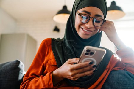 Téléchargez les photos : Jeune belle femme souriante en hijab et des lunettes assises sur le canapé avec téléphone et étançant sa tête - en image libre de droit