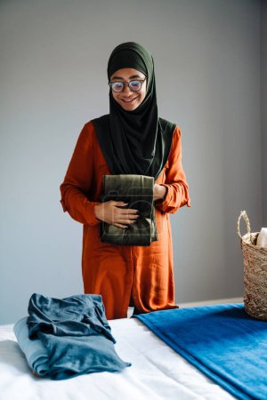 Téléchargez les photos : Jeune belle femme souriante en hijab et lunettes pliant vêtements à la maison - en image libre de droit