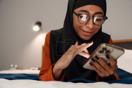 Téléchargez les photos : Jeune belle femme souriante en hijab et des lunettes couché sur le lit et en utilisant son téléphone - en image libre de droit