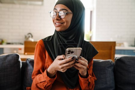 Foto de Joven hermosa mujer sonriente en hijab y gafas sentadas en el sofá con el teléfono y mirando hacia la izquierda - Imagen libre de derechos