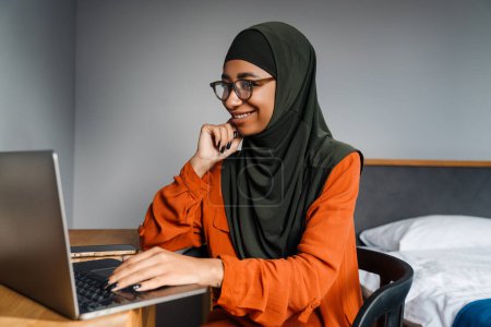 Téléchargez les photos : Jeune belle femme souriante en hijab et lunettes de travail avec ordinateur portable dans une chambre confortable à la maison - en image libre de droit