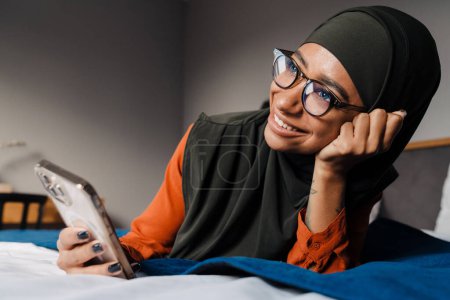 Téléchargez les photos : Jeune belle femme souriante en hijab et des lunettes couché sur le lit avec téléphone et étançant sa tête - en image libre de droit
