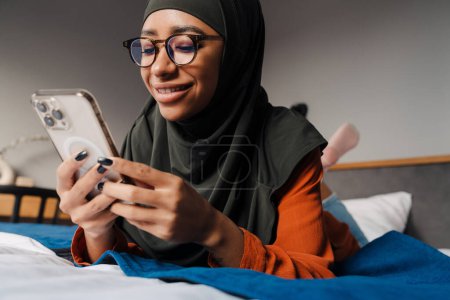 Téléchargez les photos : Jeune belle femme souriante en hijab et des lunettes couché sur le lit et regardant sur son téléphone - en image libre de droit