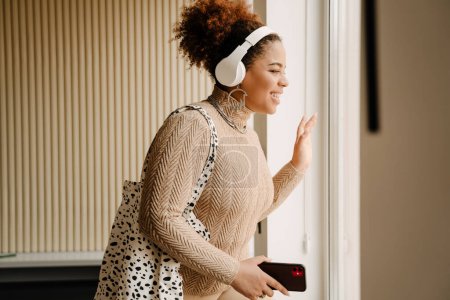 Téléchargez les photos : Vue latérale de belle femme africaine souriante dans des écouteurs avec téléphone et sac regardant par la fenêtre à la maison - en image libre de droit