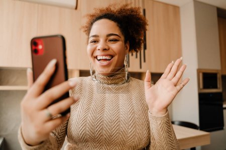 Téléchargez les photos : Jeune belle femme africaine souriante heureuse avec téléphone faisant vidéocall et agitant debout dans une cuisine confortable à la maison - en image libre de droit