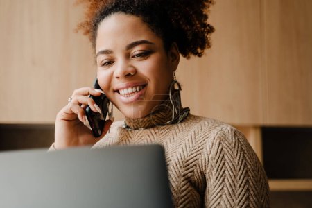 Téléchargez les photos : Jeune belle femme africaine souriante parlant sur son téléphone et regardant ordinateur portable dans sa main gros plan - en image libre de droit