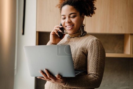 Téléchargez les photos : Jeune belle femme africaine souriante parlant sur son téléphone et regardant l'ordinateur portable dans sa main à la maison - en image libre de droit