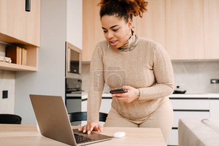 Téléchargez les photos : Jeune belle femme africaine travaillant avec ordinateur portable dans une cuisine confortable à la maison - en image libre de droit