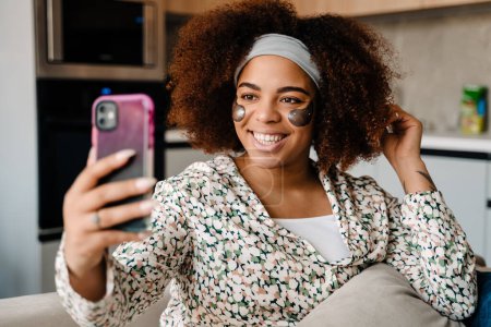 Téléchargez les photos : Jeune belle femme africaine souriante avec des patchs assis sur le canapé et prenant selfie à la maison - en image libre de droit